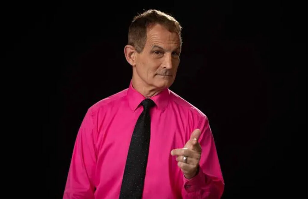 Man in a bright pink dress shirt and black tie points toward the camera against a dark background.